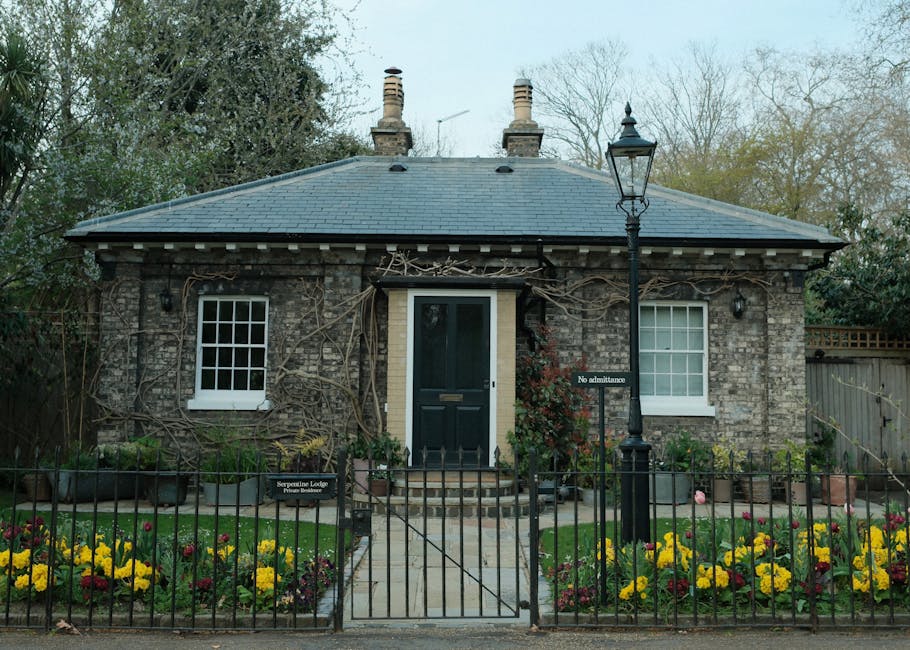 A small, single-story brick house with a gray slate roof, situated behind a black metal fence. The front garden features a paved pathway leading to the black front door, flanked by well-maintained flo