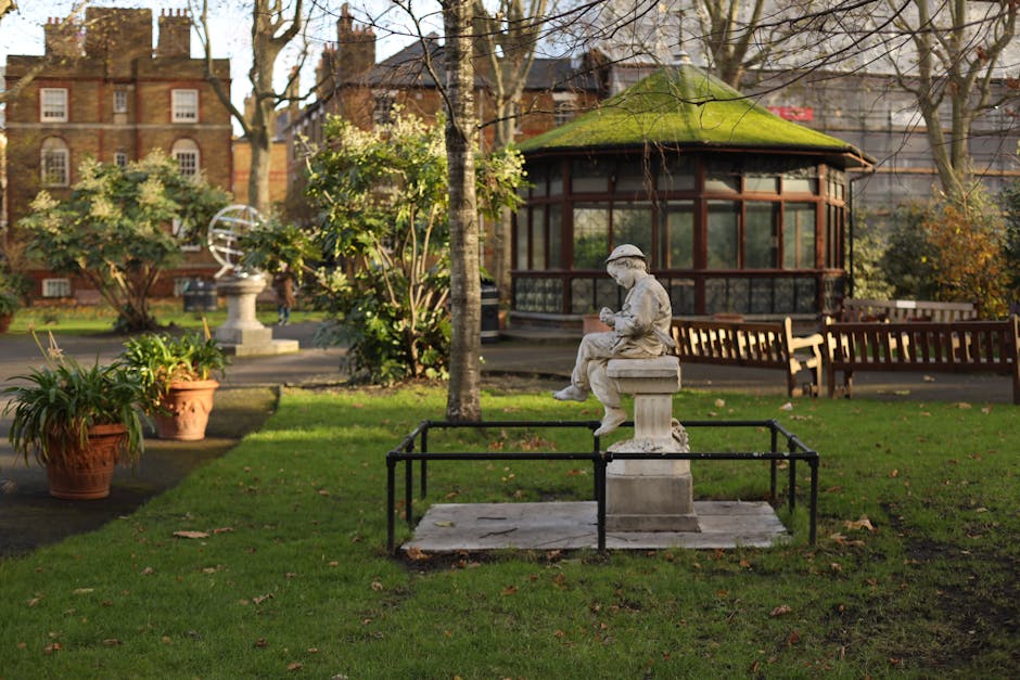 A small, single-story brick house with a gray slate roof, situated behind a black metal fence. The front garden features a paved pathway leading to the black front door, flanked by well-maintained flo