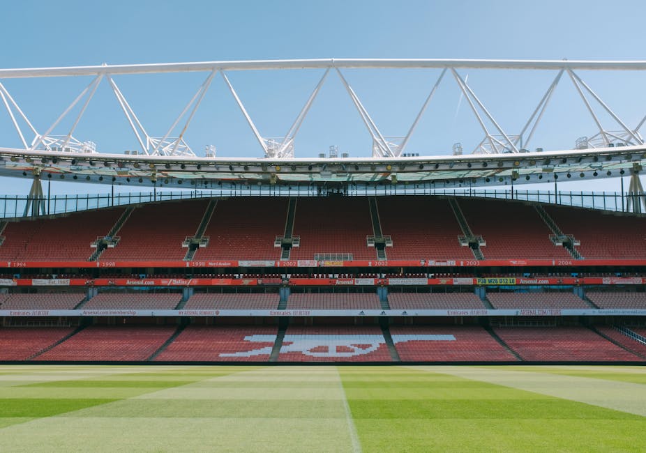 A view of the interior of the Emirates Stadium with the lush green football pitch in the foreground, featuring neatly mowed grass with visible mowing lines and a slight sheen from sunlight. Behind the